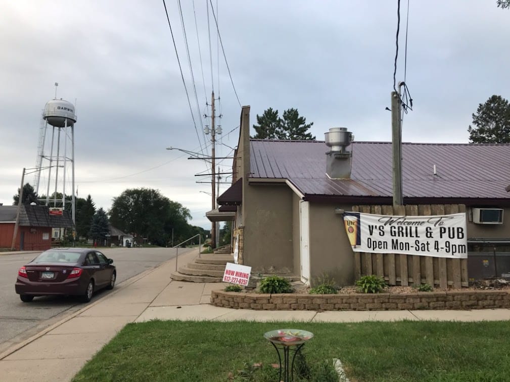 A picture of a small-town bar in profile. On the side is a sign that says "Welcome to V's Grill & Pub, Open Mon-Sat 4-9pm." A car is parked on the street, and in the background is a water tower.